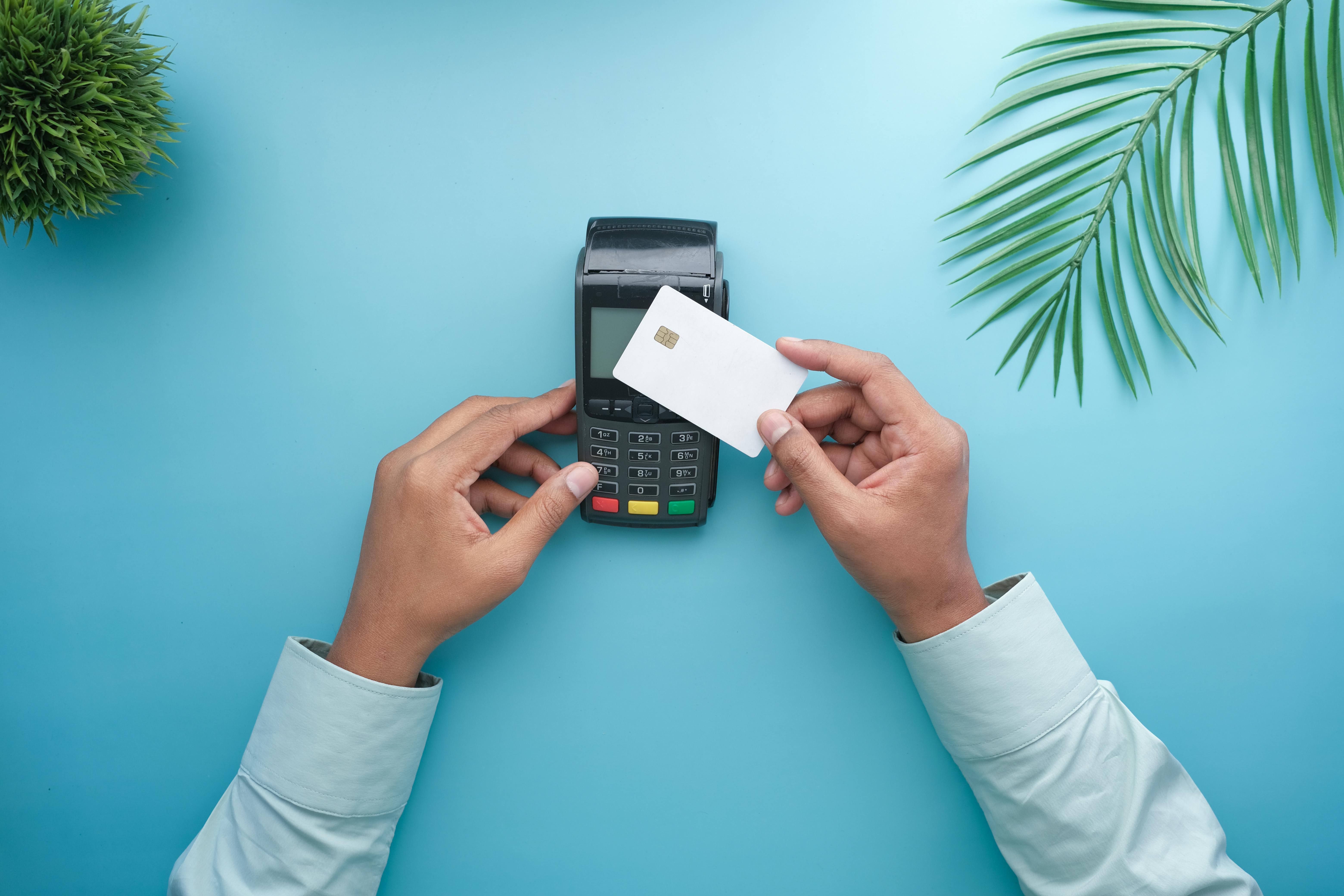 A person wearing a light blue long-sleeved shirt holds a white payment card with an EMV chip above a black POS terminal on a turquoise background, preparing to tap or insert the card for payment.