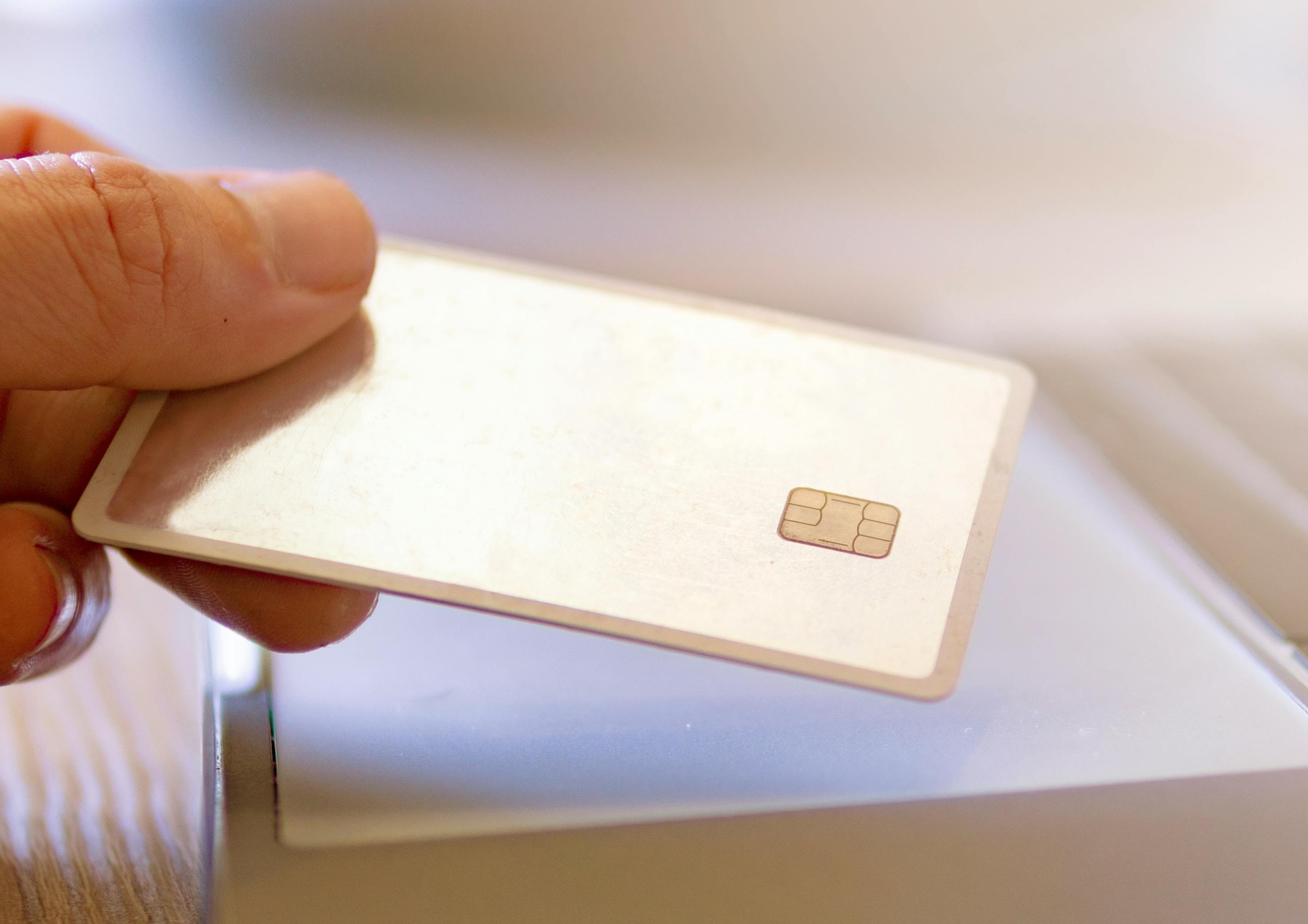 A close-up of a hand holding a white EMV chip card near a silver laptop’s keyboard, suggesting an online or contactless transaction in a well-lit indoor setting.